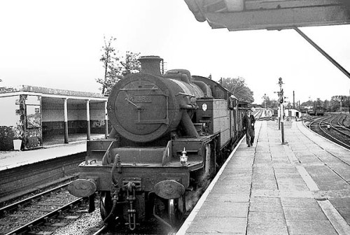 A view looking west from the island platform at Menai Bridge in August 1964. The goods train is standing at the up Afon Wen line platform which was used by passenger services travelling towards Bangor. To the left can be seen the down Afon Wen line platform. The platform that can be seen to the right served trains travelling towards Holyhead. Photo by Bevan Price